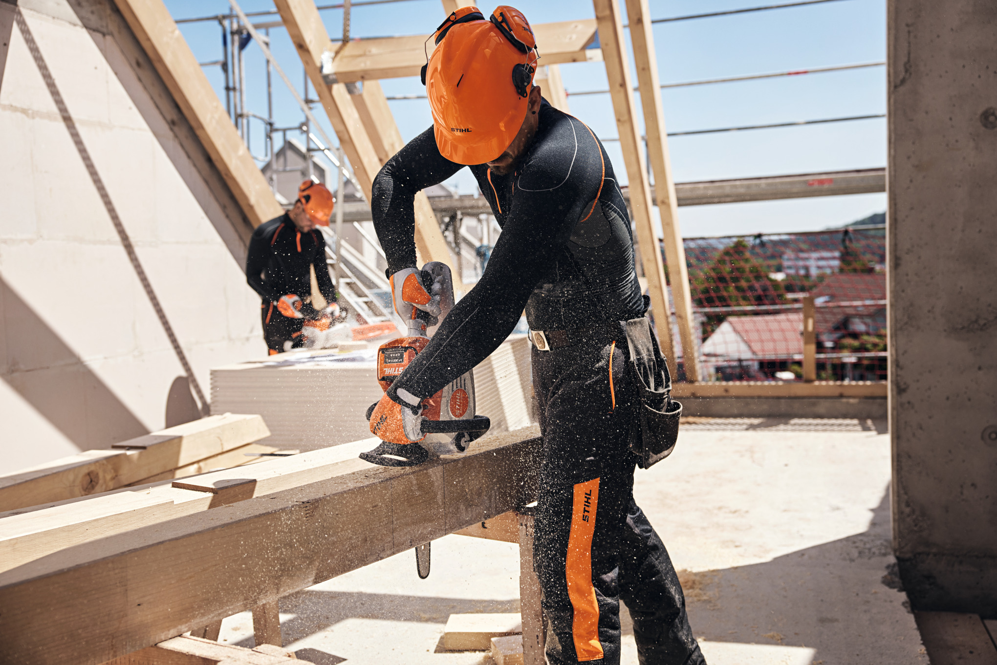 Un profesional de STIHL en una estructura del techo sierra una viga de madera con una motosierra de batería, al fondo otro trabajador maneja una motosierra a batería