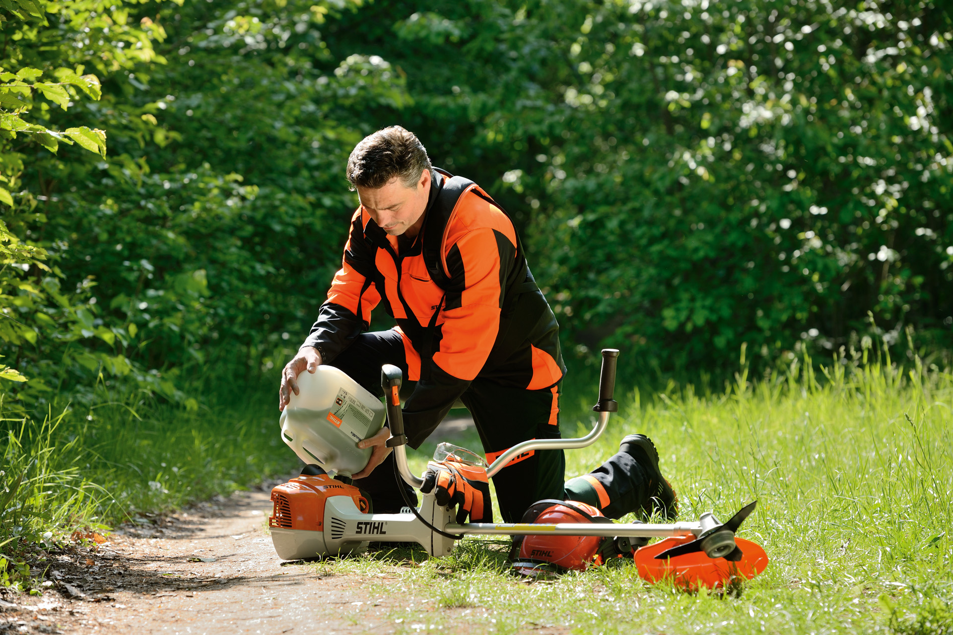 Un hombre llena el depósito de combustible de una guadaña STIHL con una garrafa con arbustos detrás de él