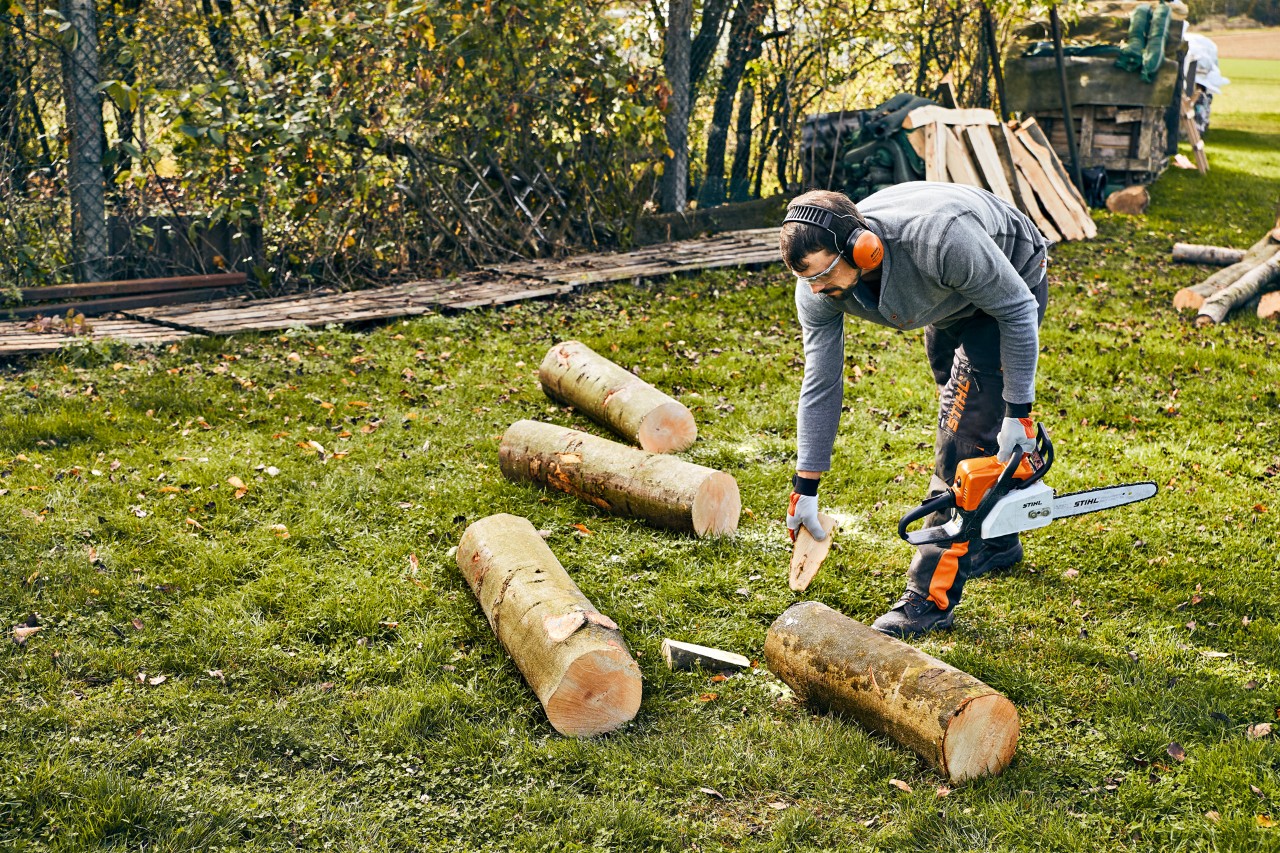 Una persona con equipamiento de protección sostiene una motosierra de gasolina STIHL MS 180 y asegura con cuñas un tronco de un metro de largo.
