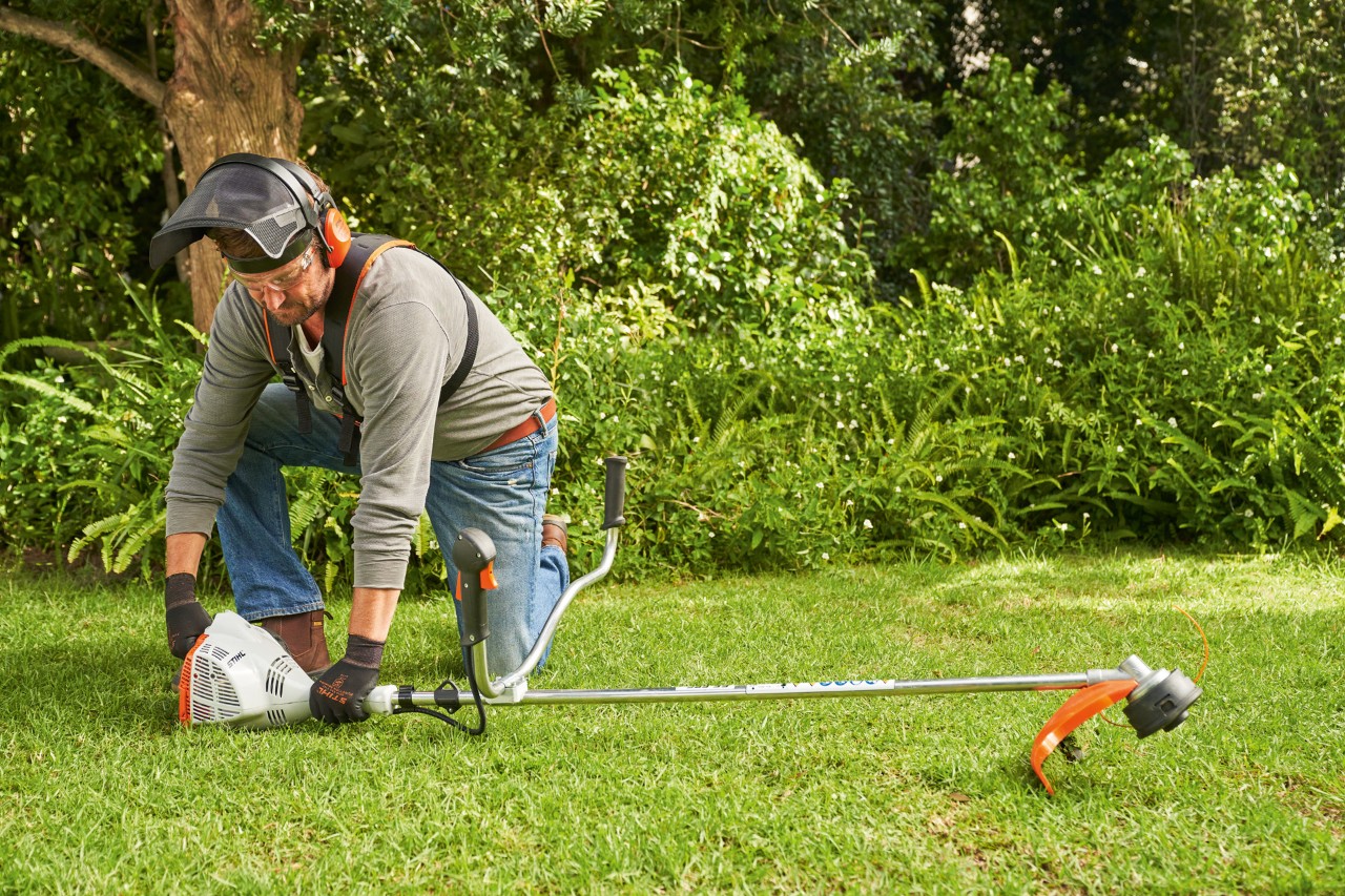 Un hombre con protección auditiva y facial arranca una desbrozadora de gasolina STIHL FS 56 R en un jardín